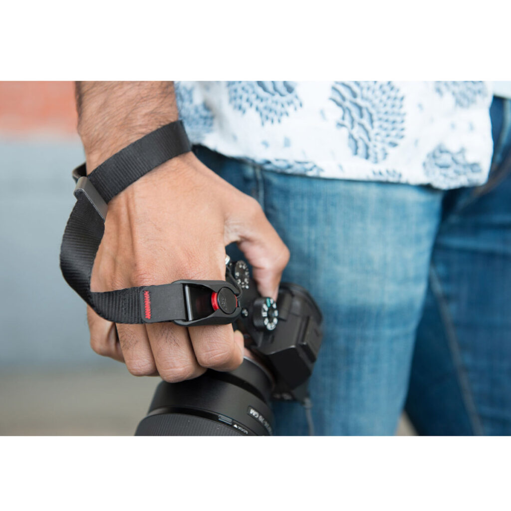Peak-Design-Cuff-Black-5 Farkkuihin ja kuvioituun paitaan pukeutunut henkilö pitää kameraa, joka on varustettu Peak Design Cuff Rannehihna - Black -rannehihnalla, valmiina ottamaan valokuvia. Keskiössä ovat hänen kätensä, musta rannehihna ja kamera.