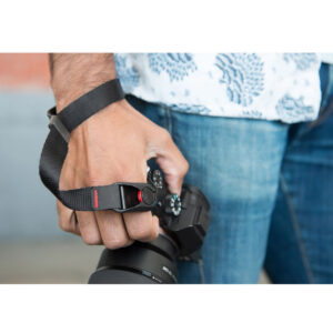 Peak-Design-Cuff-Black-5 Farkkuihin ja kuvioituun paitaan pukeutunut henkilö pitää kameraa, joka on varustettu Peak Design Cuff Rannehihna - Black -rannehihnalla, valmiina ottamaan valokuvia. Keskiössä ovat hänen kätensä, musta rannehihna ja kamera.