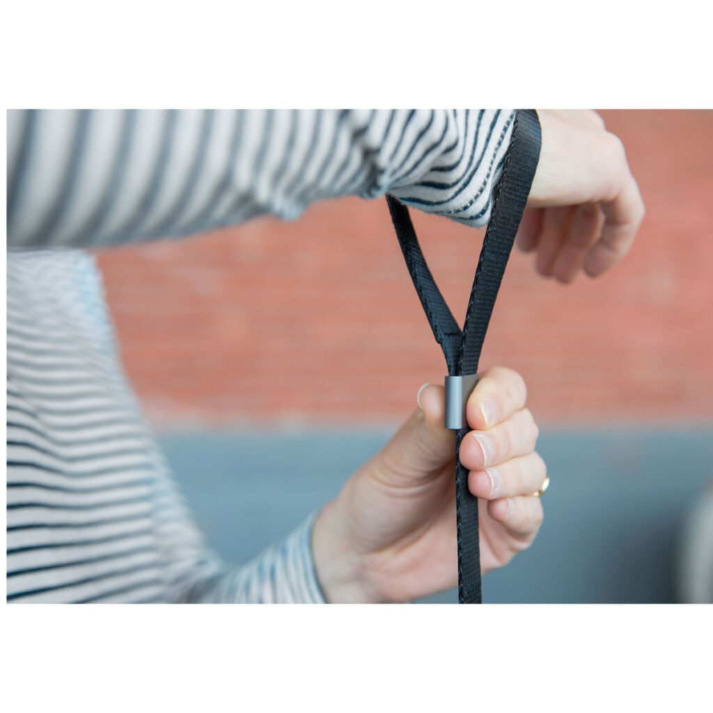 Peak-Design-Cuff-Black-6 Raidalliseen paitaan pukeutunut henkilö esittelee Peak Design Cuff Rannehihna - musta -laitteen säätöä vetämällä sen hihnan metallisilmukan läpi ja kiinnittämällä sen yhdellä kädellä epäselvää taustaa vasten.