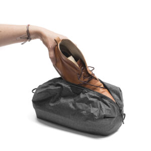 Peak-Design-Shoe-Pouch---Charcoal3 Käsi asettaa ruskean nahkakengän Peak Design Shoe Pouch - Charcoal -pussiin, jossa on valkoinen tausta.