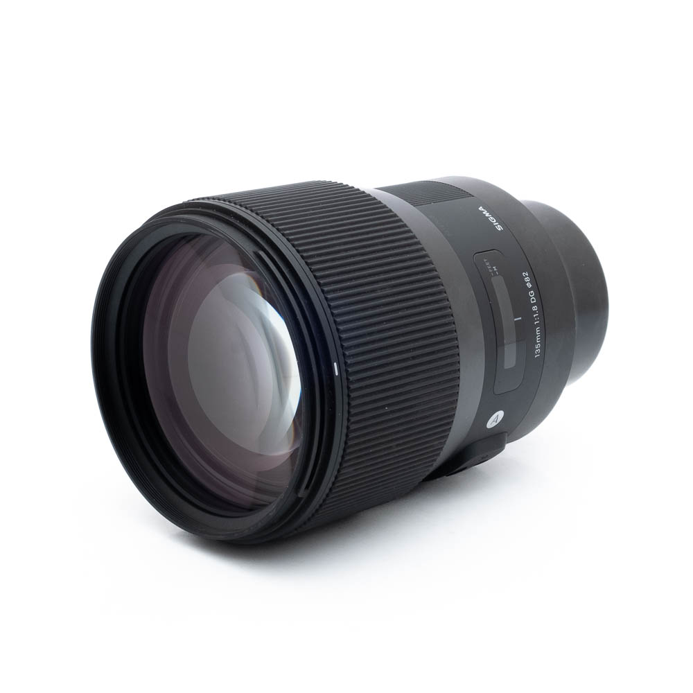 Sigma 135mm f18 Sony-2 Sigma Art 135mm f/1.8 DG Sony FE -objektiivi (sis.AVL24%) - Käytetty on esillä valkoisella taustalla, ja se on kulmailtu korostamaan sen suurta etulasia, uurrettua tarkennusrengasta ja objektiivin tynnyriä, jossa on näkyvä tuotemerkki ja mallin tiedot.