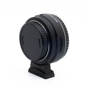 Viltrox EF-M1 Adapteri - Käytetty, musta ja suojakorkki, on esillä pienellä jalustalla valkoista taustaa vasten. Adapterissa on USB-portti ja kohdistuksen ilmaisimet.