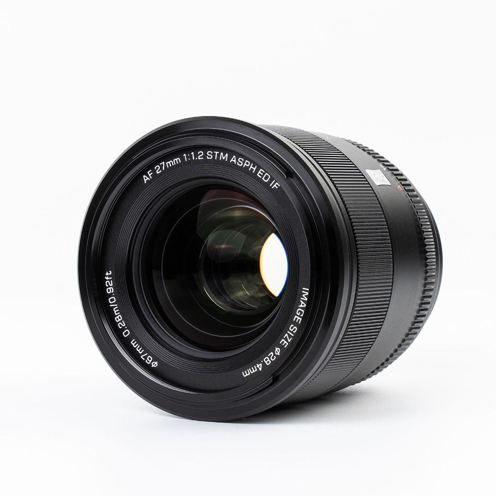 Lähikuvassa mustasta Viltrox AF PRO 27mm f/1.2 -objektiivi, Fuji X -objektiivista näkyvät sen lasielementit ja yksityiskohtainen teksti AF 27mm 1:1.2 STM ASPH ED IF, joka korostaa teknisiä tietoja valkoista taustaa vasten.