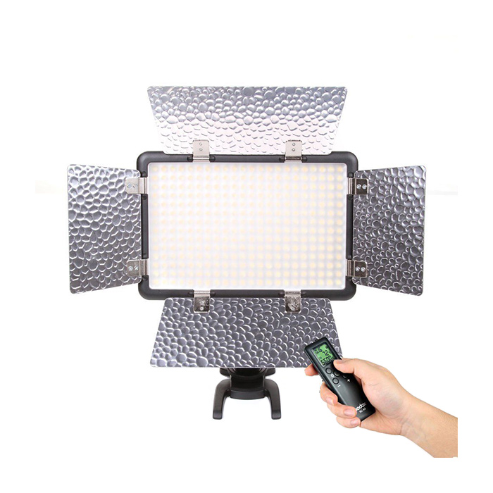 1446963156_452 Godox LED 308C II suorakulmainen studiovalo, jossa on neljä säädettävää hopeista lato-ovea ja kädessä pidetty digitaalinen kaukosäädin valkoista taustaa vasten.