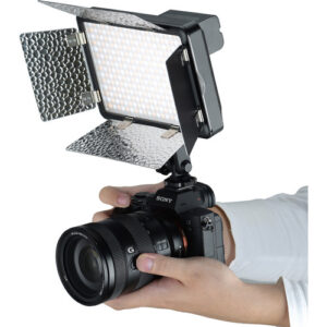 1579519632_537 Henkilö pitää kädessään Sony-kameraa, jossa on suuri objektiivi ja suorakulmainen Godox Led LF308BI -videovalaisin, jonka päälle on asennettu heijastavat lato-ovet.