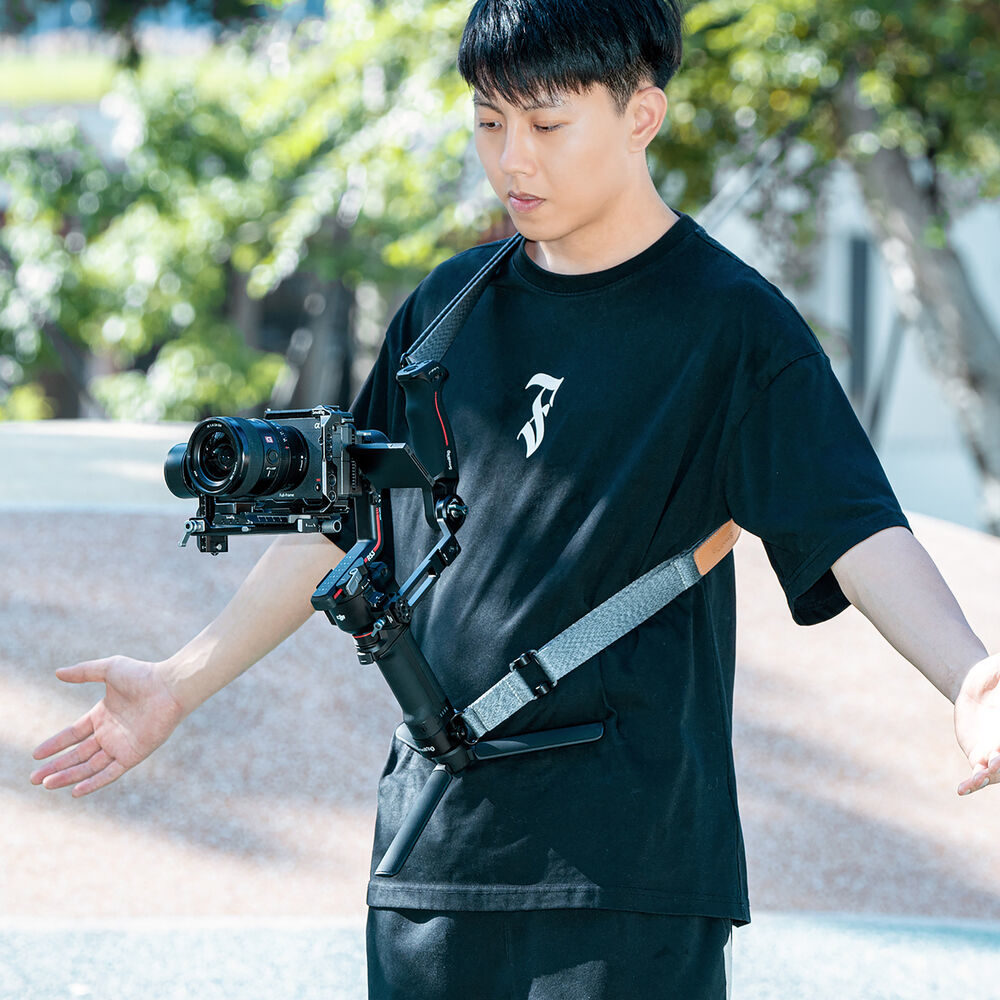 1698674746_664 Mustaan t-paitaan pukeutunut henkilö käyttää SmallRig 4118B Weight Reducing Shoulder Strap for DJI RS 3/RS 3 Pro/RS 2/RSC 2 -olkahihnaa tukemaan kameran vakaajaa kuvatessaan ulkona aurinkoisena päivänä, kun taustalla on puita ja vehreyttä.