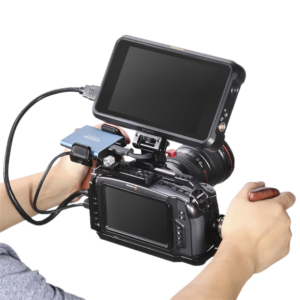 800q-05_114208 Henkilö pitää kädessään ammattikäyttöön tarkoitettua videokameralaitetta, jossa on SmallRig 2117 Right Side Wooden Grip with NATO Mount, sekä suurta ulkoista monitoria, objektiivia ja lisävarusteita, kaikki kuvattuna valkoista taustaa vasten.
