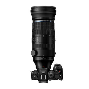 OM_150-600mm_004 OM System M.Zuiko Digital ED 150-600mm f/5.0-6.3 IS -objektiivi on kiinnitetty digitaalikameraan ylhäältä katsottuna valkoisella taustalla. Mustassa teleobjektiivissa on 150-600 mm:n polttoväli, ja siihen sisältyy 600 euron käteispalautustarjous.