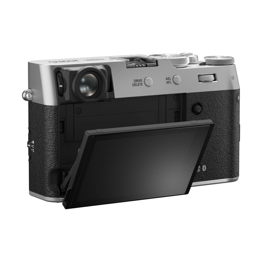 fuji_x100vi_hopea_01 Fujifilm X100VI -kompaktikamera, hopea näkyy takaa sen hopea ja musta runko. LCD-näyttö on käännetty ulos ja kallistettu alaspäin, jolloin kameran hallintalaitteet ja kuvioitu kahva paljastuvat.