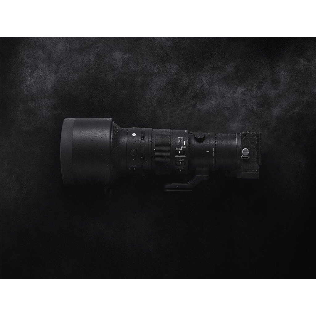 sigma_500mm_dn_01 Sigma 500mm f/5.6 DG DN OS Sports -objektiivi Sonylle on esillä vaakasuorassa linssinsuojuksen kanssa tummaa, kuvioitua taustaa vasten. Sen mustalla pinnalla olevat vesipisarat korostavat sen säänkestävää muotoilua.