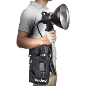 1455605238_315 A man in a grey polo shirt is holding a Godox AD-H1200B Portable Flash Head Bowens flashlight with a large reflector, connected by cable to its control unit, which is carried in a black shoulder bag.