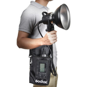 1455605238_315 Harmaaseen poolopaitaan pukeutunut henkilö pitää kädessään Godox AD-H1200B Portable Flash Head Bowens -salamalaitetta, jossa on suuri heijastin ja joka on liitetty kaapelilla sen ohjausyksikköön, jota kannetaan mustassa olkalaukussa.