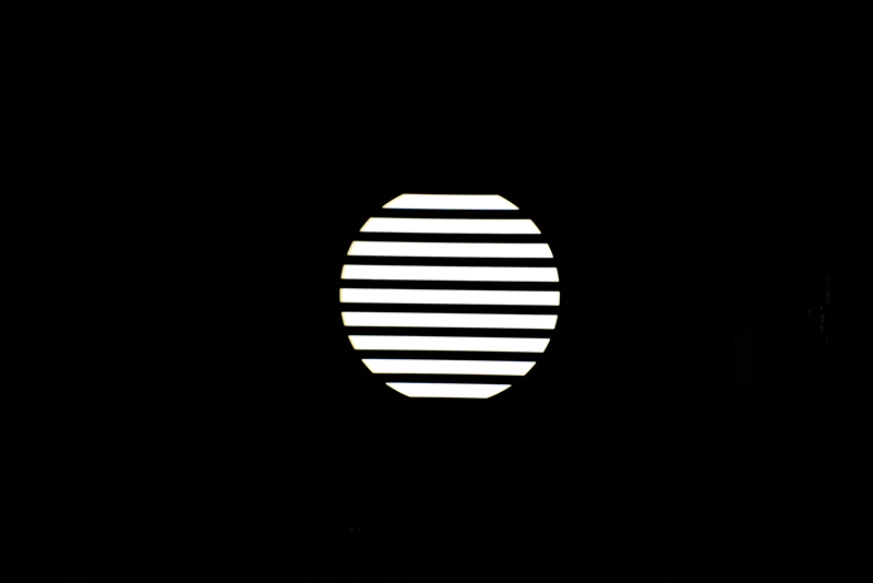 800q-04_117077 NANLITE GOBO Set 1 for PJ-FMM heijastaa kirkasta valkoista valoa vaakasuorilla säleillä varustetun pyöreän ikkunan läpi, jolloin pyöreän muodon sisällä olevat raidalliset kuviot näkyvät pimeässä huoneessa.