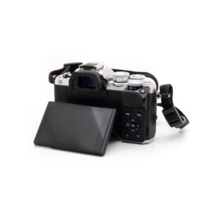 Olympus OM-D E-M10 Mark III (SC 8200) - Käytetty on esitetty valkoisella pohjalla, ja sen nestekidenäyttö on käännetty ulospäin, jolloin kameran painikkeet ja säätimet tulevat näkyviin. Kiinnitetty hihna lepää laitteen vieressä.