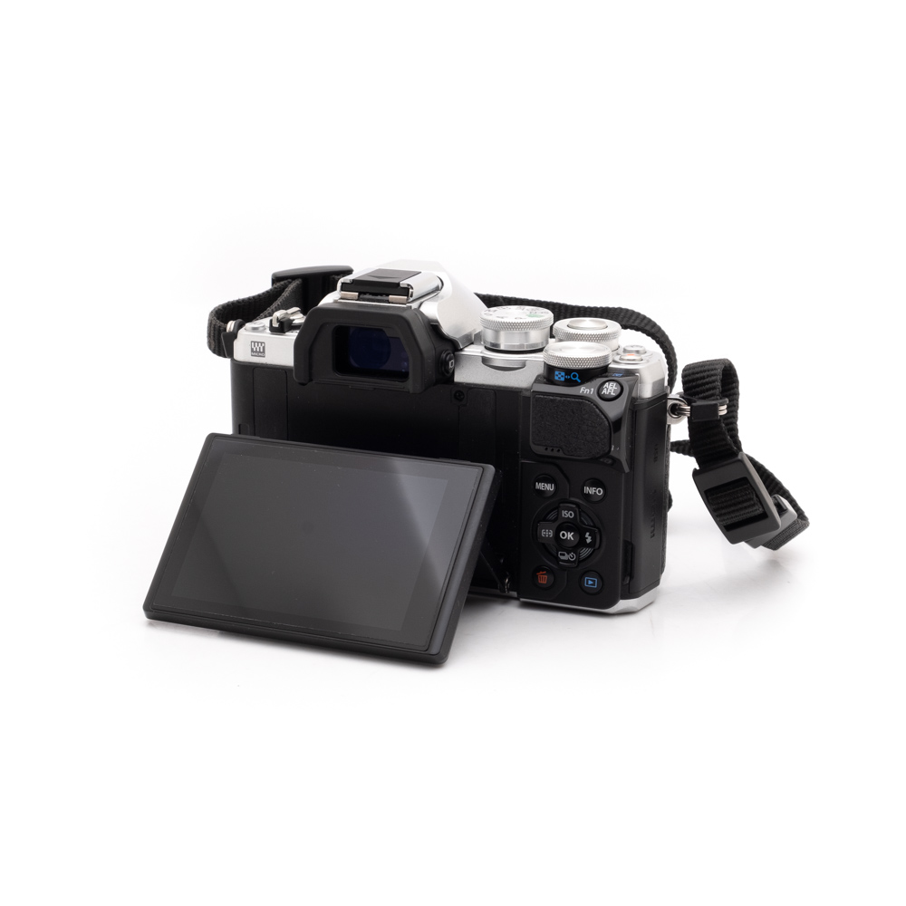 Olympus OM-D E-M10 Mark III (SC 8200) - Käytetty on esitetty valkoisella pohjalla, ja sen nestekidenäyttö on käännetty ulospäin, jolloin kameran painikkeet ja säätimet tulevat näkyviin. Kiinnitetty hihna lepää laitteen vieressä.