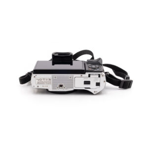 Olympus OM-D E-M10 Mark III (SC 8200) - Käytetty, musta-hopea digikamera, jossa on ulos käännettävä näyttö, ohjauspainikkeet, käsihihna ja useita portteja ja tarroja pohjassa, on kuvassa selällään.