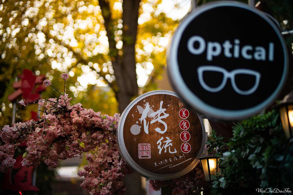 1545211561_071 Viltrox MF 85mm f/1.8 PFU RBMH -objektiivilla Sonylle kuvattu kiinalaisilla kirjaimilla koristeltu puinen kyltti on vaaleanpunaisten kukkien kehystämä, kun taas pyöreä optinen kyltti silmälaseineen erottuu auringonvalossa keltaisine lehvästöineen puiden edessä.
