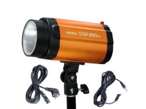 1646831718_976 Oranssi Godox Mini Pioneer 250W Softbox Umbrella Duo Kit -stroboskooppivalaisin on asennettu jalustalle, ja sen vieressä on kaksi mustaa virtajohtoa valkoisella taustalla.