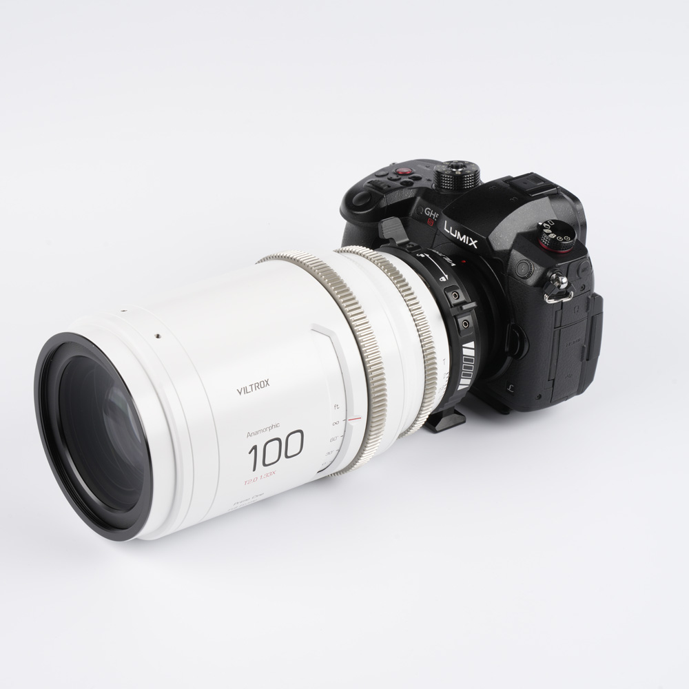 1709809410_923 Musta Panasonic Lumix GH6 -kamera, jossa on suuri valkoinen Viltrox 100mm T2.0 -kino-objektiivi, on kiinnitetty Viltrox PL - MFT -sovittimella, ja kaikki on asetettu valkoista taustaa vasten.