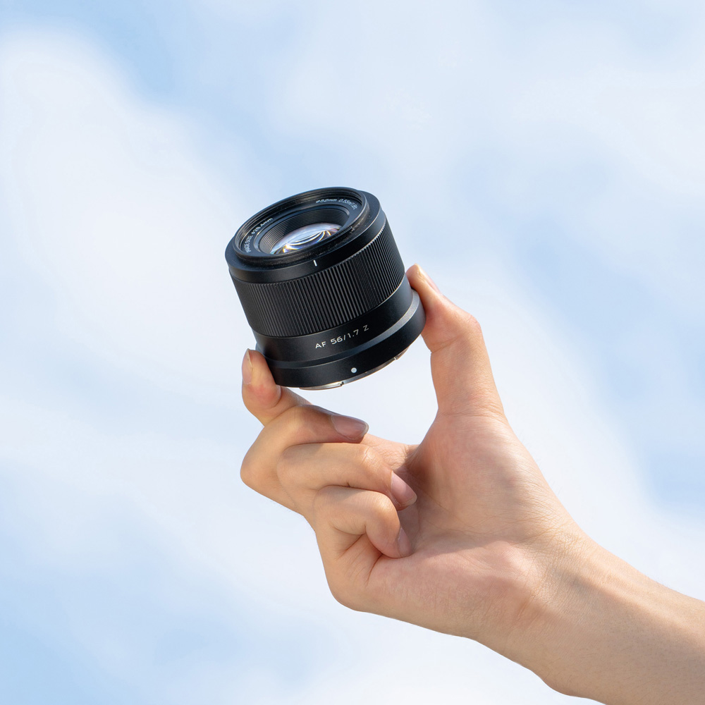 1712655719_627 Käsi pitää ylhäällä Viltrox AF 56mm f/1.7 -objektiivi, Nikon Z -objektiivia kirkkaan sinistä taivasta vasten, jonka taustalla on pehmeät valkoiset pilvet.