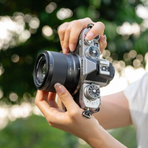1712655720_706 Henkilö pitelee hopean ja mustan väristä Viltrox AF 56mm f/1.7 -objektiivi for Nikon Z -kameraa vaakatasossa ulkona, vaaleat kynnet näkyvät ja taustalla on sumea vihreä tausta.