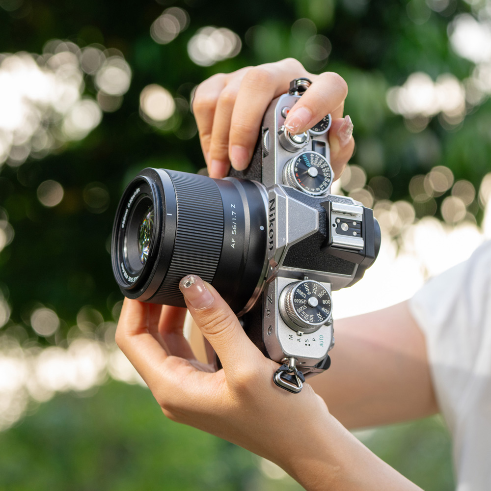 1712655720_706 Henkilö pitelee hopean ja mustan väristä Viltrox AF 56mm f/1.7 -objektiivi for Nikon Z -kameraa vaakatasossa ulkona, vaaleat kynnet näkyvät ja taustalla on sumea vihreä tausta.