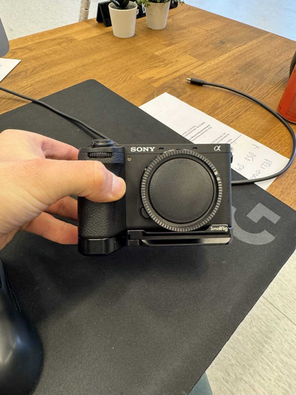 IMG_1852 Käsi pitää mustaa Sony A6700 (Kunto K5) + kahva - Käytetty -kamerarunkoa ilman objektiivia tummalla hiirimatolla puupöydän päällä. Taustalla näkyy papereita, kynä ja pieniä ruukkukasveja.