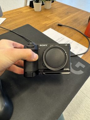 Käsi pitää mustaa Sony A6700 (Kunto K5) + kahva - Käytetty -kamerarunkoa ilman objektiivia tummalla hiirimatolla puupöydän päällä. Taustalla näkyy papereita, kynä ja pieniä ruukkukasveja.