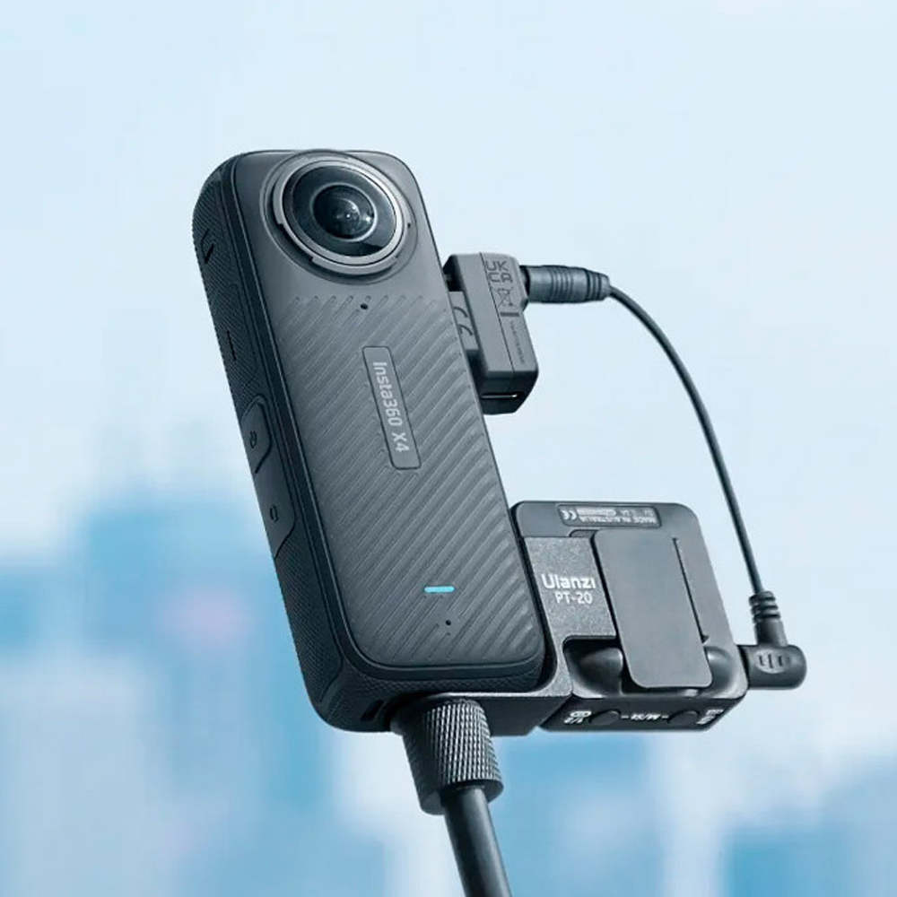 Insta360_X4_044 Close-up of the Insta360 X4 Mic Adapter connected to a black 360-degree action camera mounted on a tripod and equipped with cables. The blurred background shows a blue-tinted cityscape.