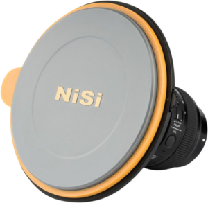 Lähikuva kameran objektiivista, johon on asennettu NiSi Lens Cap for S5/S6 Holder -objektiivin suojus, jossa näkyy sen pyöreä harmaa ja oranssi muotoilu, ja osa objektiivista ja tarkennuskytkimistä on näkyvissä tuotemerkillä varustetun suojuksen takana.