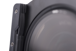 800q-02_113503 Lähikuva Nisi Filter Holder for Laowa 12mm f/2.8 -suodattimen pidikkeestä kiinnitettynä kameran objektiiviin, korostaen sen mustaa muotoilua, näkyviä ruuveja ja reunoja vaaleaa, neutraalia taustaa vasten.