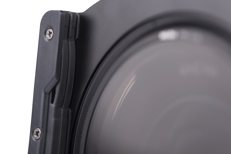 800q-02_113503 Lähikuva Nisi Filter Holder for Laowa 12mm f/2.8 -suodattimen pidikkeestä kiinnitettynä kameran objektiiviin, korostaen sen mustaa muotoilua, näkyviä ruuveja ja reunoja vaaleaa, neutraalia taustaa vasten.