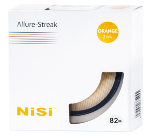 800q-02_121024 Valkoinen laatikko, jossa lukee "Nisi Filter Allure Streak Orange 2mm 82mm", näyttää oranssin kamerasuodattimen oikeassa alakulmassa olevan kirkkaan ikkunan läpi. Pakkaus on koristeltu värillisillä viivoilla ja tekstillä.