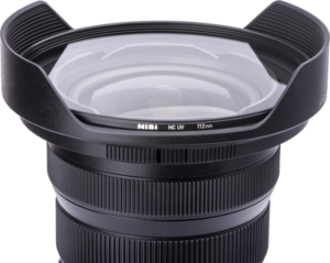 800q-03_114918 Lähikuva mustasta kameran objektiivista, jossa on laajakulmahuppu ja NiSi NC UV 112mm Filter For Nikon Z 14-24mm f/2.8 -UV suodin -etiketti suodattimessa valkoista taustaa vasten.