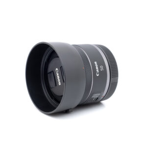 Canon RF 50mm f/1.8 STM - Käytetty -objektiivi, johon on kiinnitetty vastavalosuoja, kyljellään valkoista taustaa vasten.