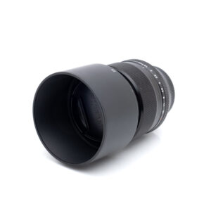 Fujinon XF 56mm f/1.2 R - Käytetty objektiivi mustana, johon on kiinnitetty suuri huppu, makaa kyljellään valkoista taustaa vasten.