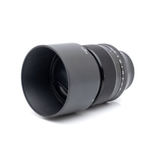 Fujinon XF 56mm f/1.2 R - Käytetty objektiivi, johon on kiinnitetty vastavalosuoja, on suunnattu valkoiselle taustalle, mikä korostaa sen mustaa pintaa sekä tarkennusrengasta ja aukkorenkaita.