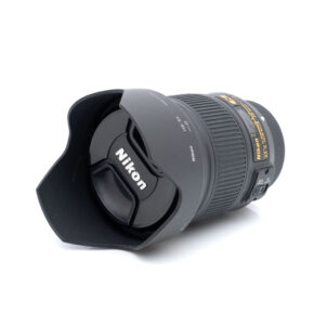 Nikkor 28mm f1.8 G-1 Nikon AF-S Nikkor 28mm f/1.8 G - Käytetty -objektiivi, johon on kiinnitetty vastavalosuoja ja suojus, makaa kyljellään valkoista taustaa vasten, ja Nikonin logo näkyy objektiivin suojuksessa.