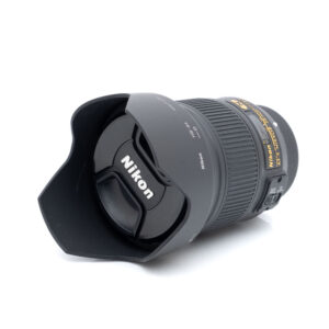 Nikkor 28mm f1.8 G-1 Nikon AF-S Nikkor 28mm f/1.8 G - Käytetty -objektiivi, johon on kiinnitetty vastavalosuoja ja suojus, makaa kyljellään valkoista taustaa vasten, ja Nikonin logo näkyy objektiivin suojuksessa.