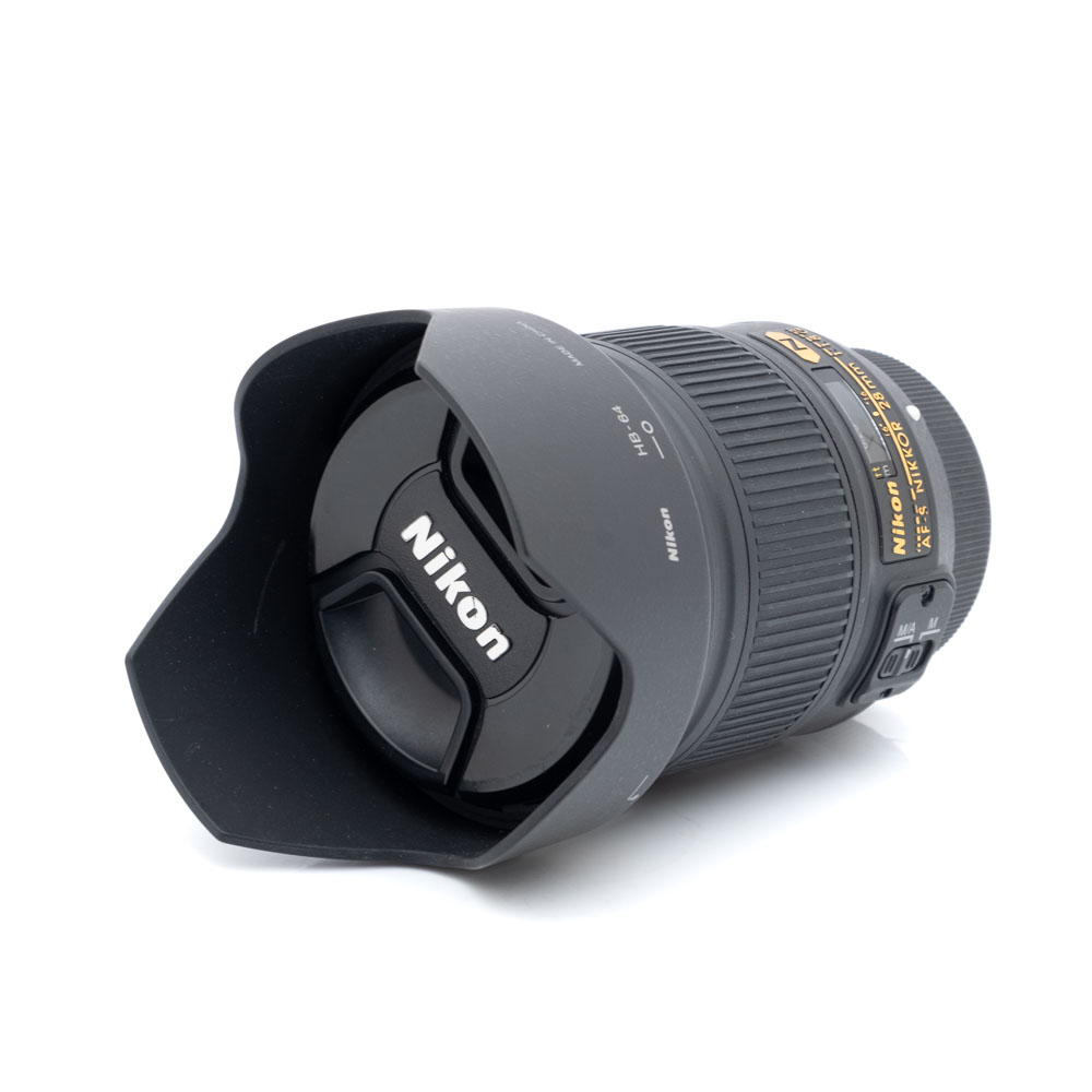 Nikkor 28mm f1.8 G-1 Nikon AF-S Nikkor 28mm f/1.8 G - Käytetty -objektiivi, johon on kiinnitetty vastavalosuoja ja suojus, makaa kyljellään valkoista taustaa vasten, ja Nikonin logo näkyy objektiivin suojuksessa.
