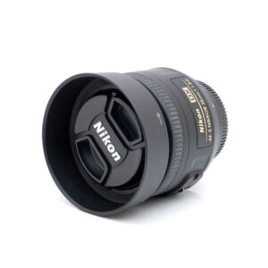 Nikon AF-S Nikkor 35mm f/1.8G DX - Käytetty -objektiivi, johon on kiinnitetty vastavalosuoja ja suojus, näkyy kulmassa valkoisella taustalla. Sivussa näkyvät kultaiset Nikon- ja DX-tarrat sekä tarkennuksen ja objektiivin tiedot.