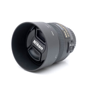 Nikkor 50mm f-1.4 G-1 Nikon AF-S Nikkor 50mm f/1.4 G (Kunto K5) - Käytetty objektiivi vastavalosuojalla ja suojuksella, jossa näkyy Nikonin logo ja tarkennustilan kytkimet sivussa, kuvattuna valkoista taustaa vasten.