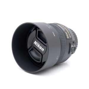 Nikkor 50mm f-1.4 G-1 Nikon AF-S Nikkor 50mm f/1.4 G (Kunto K5) - Käytetty objektiivi vastavalosuojalla ja suojuksella, jossa näkyy Nikonin logo ja tarkennustilan kytkimet sivussa, kuvattuna valkoista taustaa vasten.