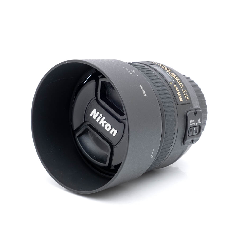 Nikkor 50mm f-1.4 G-1 Nikon AF-S Nikkor 50mm f/1.4 G (Kunto K5) - Käytetty objektiivi vastavalosuojalla ja suojuksella, jossa näkyy Nikonin logo ja tarkennustilan kytkimet sivussa, kuvattuna valkoista taustaa vasten.