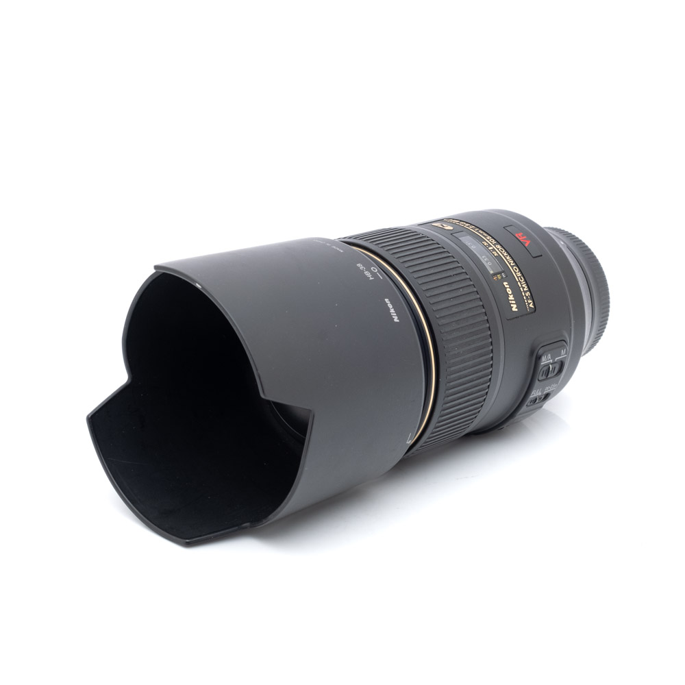 Nikkor AF-S 105mm f2.8-1 Nikon AF-S Micro-Nikkor 105mm f/2.8G VR ED - Käytetty -objektiivi, johon on kiinnitetty terälehden muotoinen huppu, valkoista taustaa vasten.