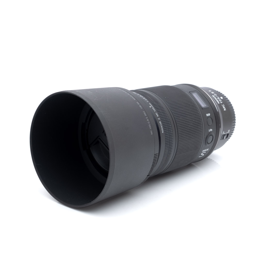 Nikkor MC 105mm f2.8 S-1 Nikon Nikkor Z MC 105mm f/2.8 VR S (Kunto K5) - Käytetty teleobjektiivi, johon on kiinnitetty vastavalosuoja, valkoisella pinnalla ja katsojaa kohti suunnattuna.