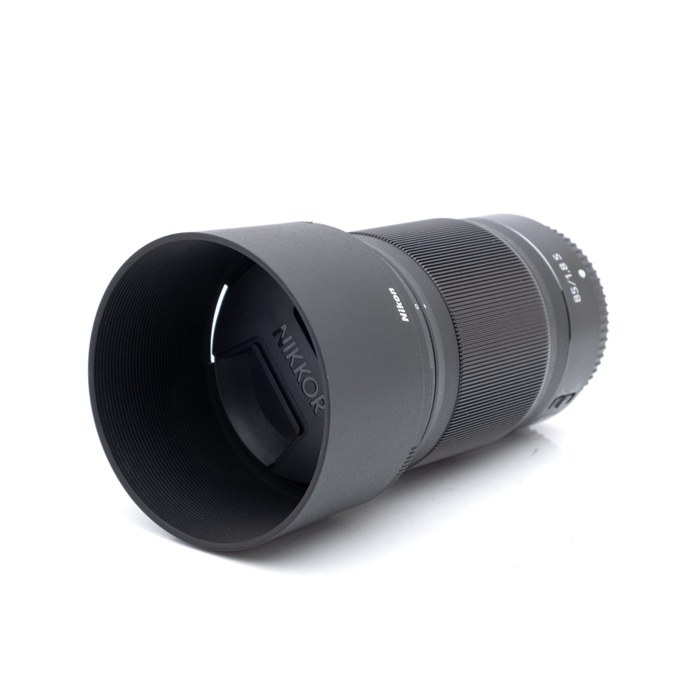Nikkor Z 85mm f1.8 S-1 Musta Nikon Nikkor Z 85mm f/1.8 S (Kunto K5) -objektiivi, jossa on vastavalosuoja ja suojus, makaa kyljellään valkoista taustaa vasten. Tarkennus- ja zoomausrenkaat sekä tekniset merkinnät ovat näkyvissä objektiivin rungossa.