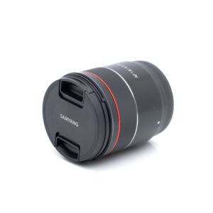 Samyang 18mm f2.8 Sony-1 Samyang AF 18mm f/2.8 Sony FE - Käytetty kameran objektiivi, jossa on objektiivin suojus, musta runko ja punainen rengas, makaa kyljellään valkoista taustaa vasten.