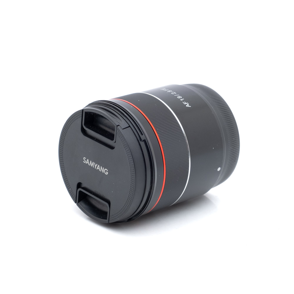 Samyang 18mm f2.8 Sony-1 Samyang AF 18mm f/2.8 Sony FE - Käytetty kameran objektiivi, jossa on objektiivin suojus, musta runko ja punainen rengas, makaa kyljellään valkoista taustaa vasten.