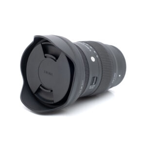 Sigma 16-28mm f2.8 Sony-1 Sigma 16-28mm f/2.8 DG DN C Sony E -objektiivi (käytetty, kunto K5, 24 kk takuu, sis. alv 24%), jossa on vastavalosuoja ja suojus, makaa kyljellään valkoista taustaa vasten.
